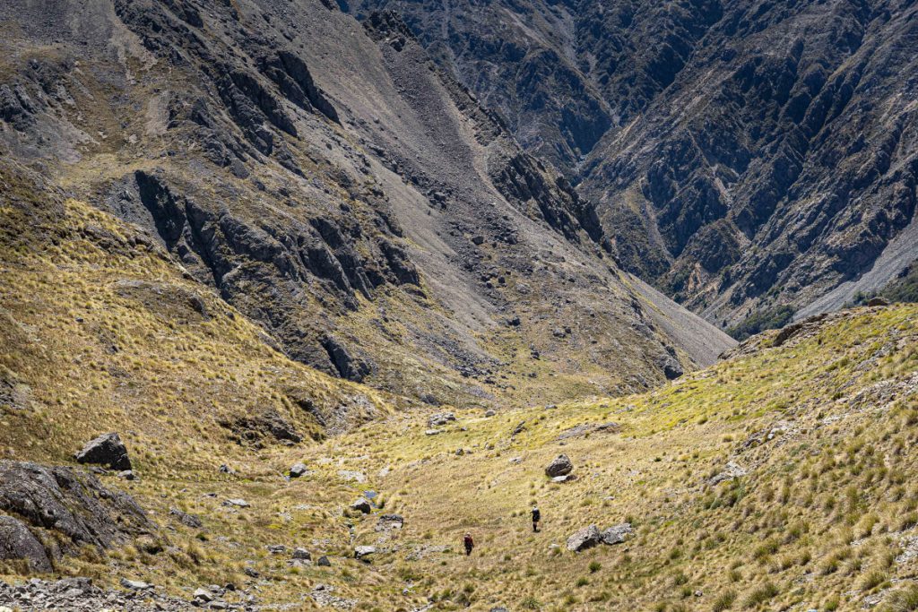 View down a valley with two small trampers visible and steep rock and scree faces