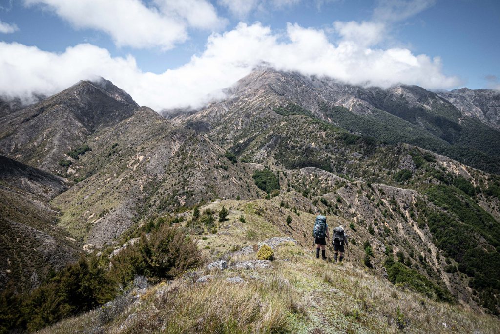 Two trampers walk along a ridge with mountains in the background