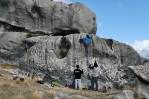 photo of a woman climbing on a large rock boulder with 2 people spotting her